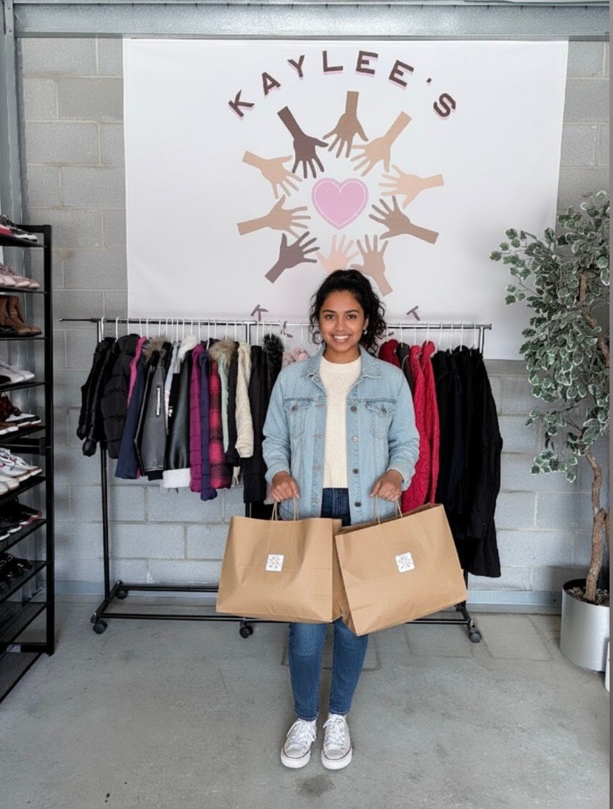 Woman holding two cardboard boxes in front of a clothing rack and “KAYLEE’S” sign.