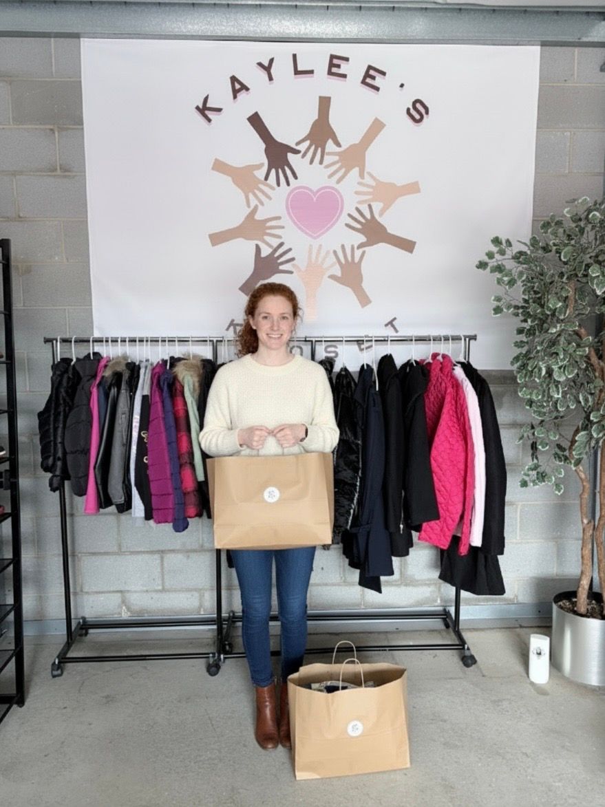 Woman holding a box in front of a clothing rack with “Kaylee’s” sign and folded clothes