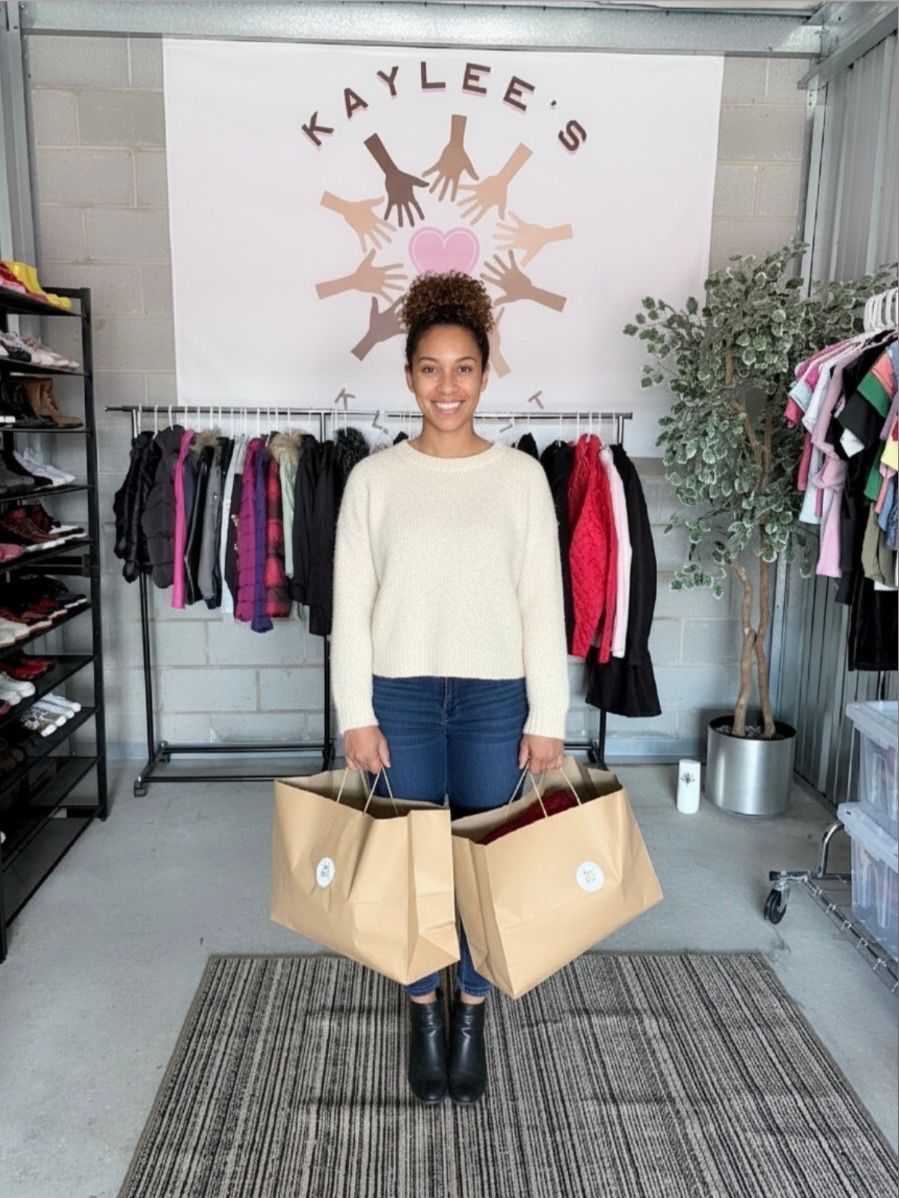 Woman standing in a boutique holding two tan shopping bags, with clothing racks behind her