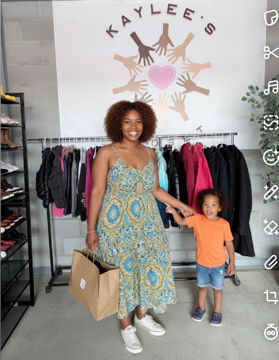 Woman and toddler holding hands in a boutique, with clothing racks and a “Kaylee’s” wall logo behind them.
