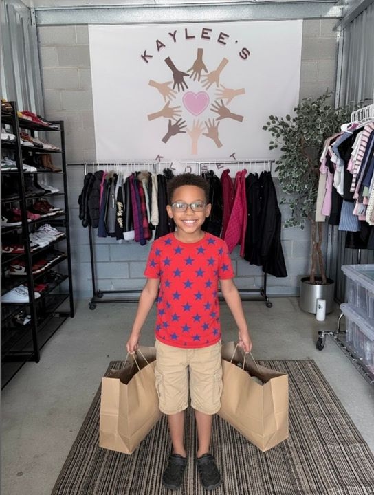 Two people in a clothing boutique holding tan shopping bags, with racks of clothes and a “Kaylee’s” sign behind them.
