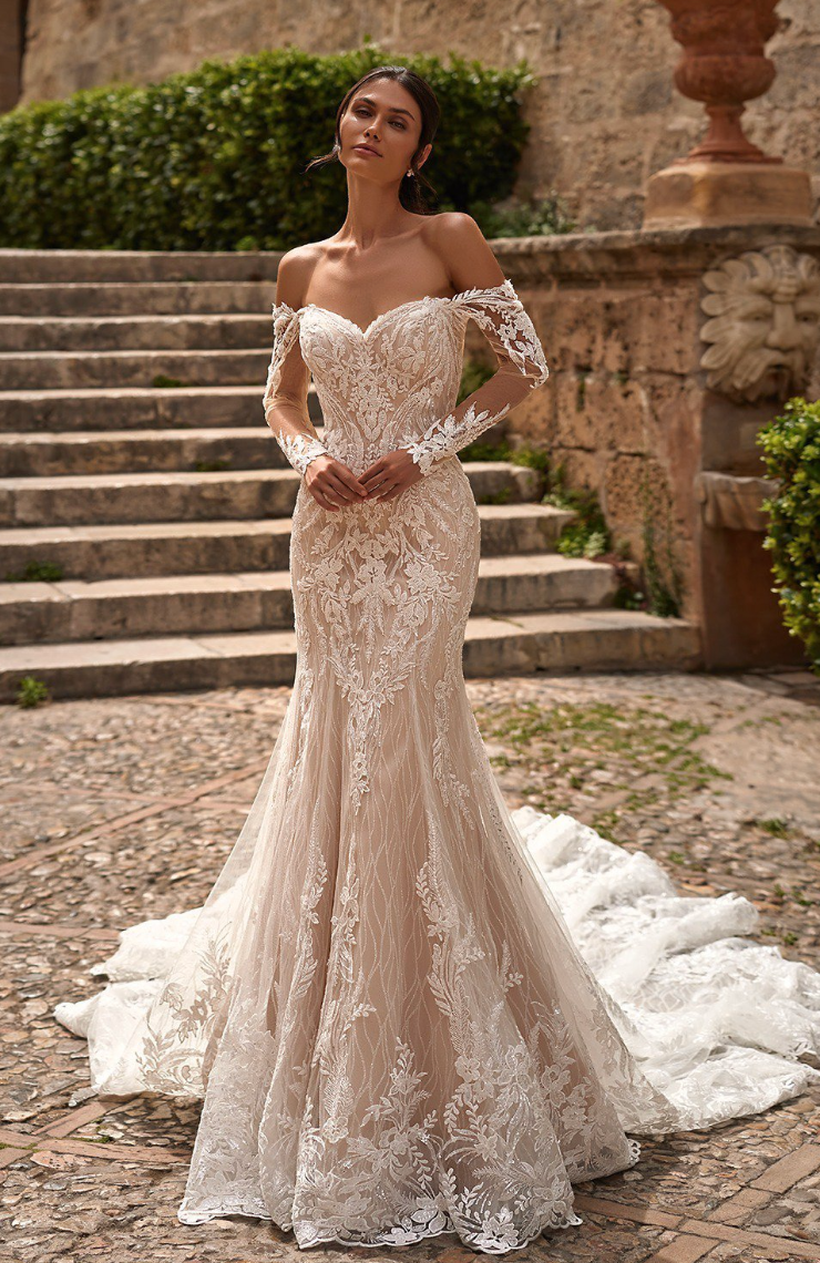 Woman in a white lace off-the-shoulder wedding dress with long sleeves, standing on stone steps, train on ground.