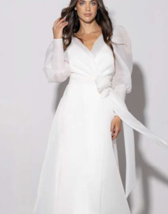 A woman in a white dress with long sleeves is standing in front of a white wall.