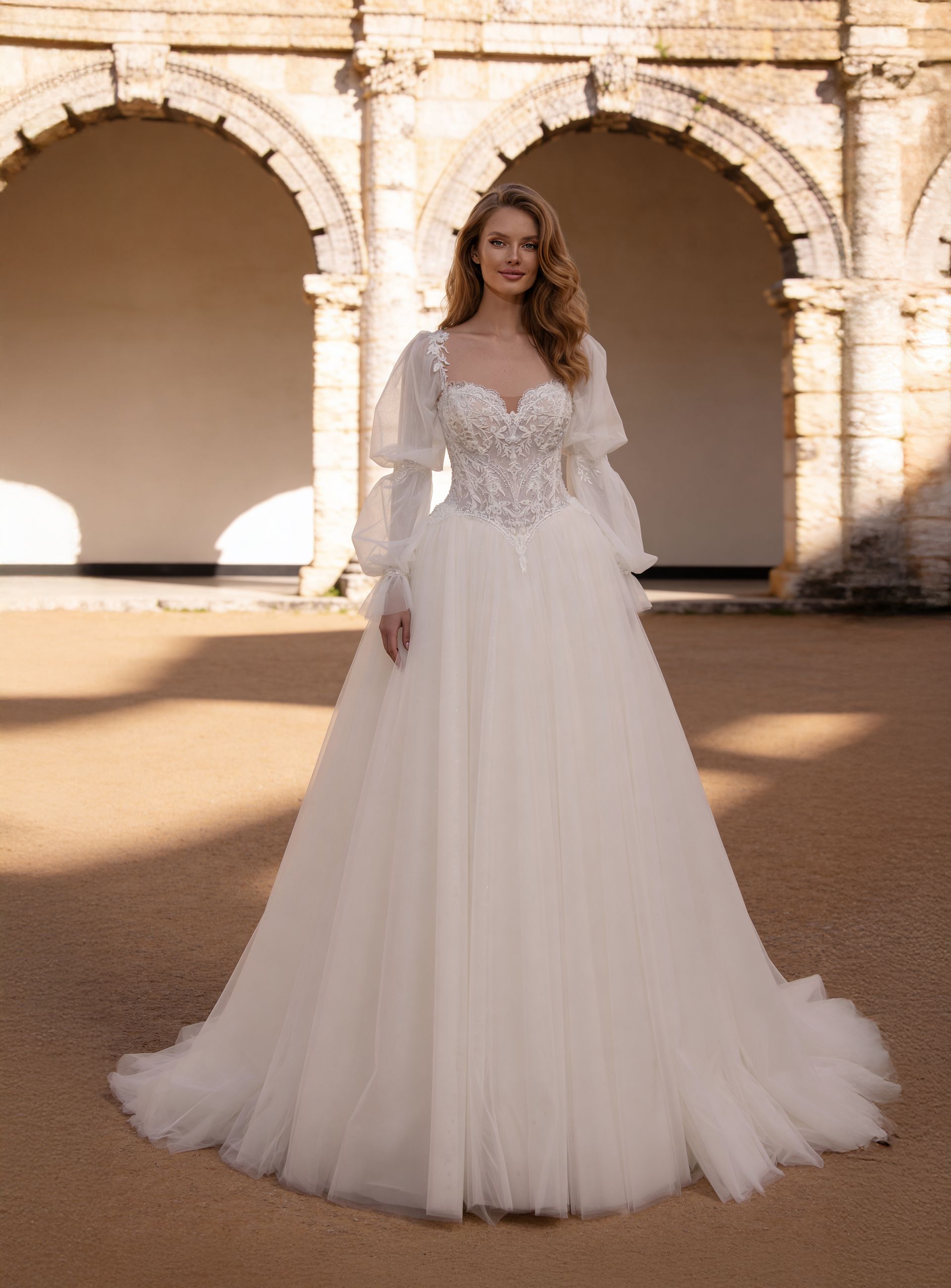 Woman in a white lace bridal gown standing under sunlit stone arches