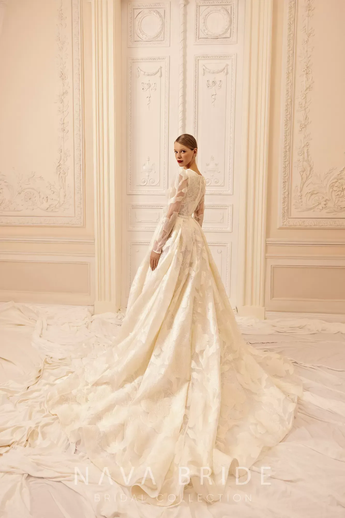 Woman in an embellished ivory wedding gown with long train, standing indoors in front of ornate doors.