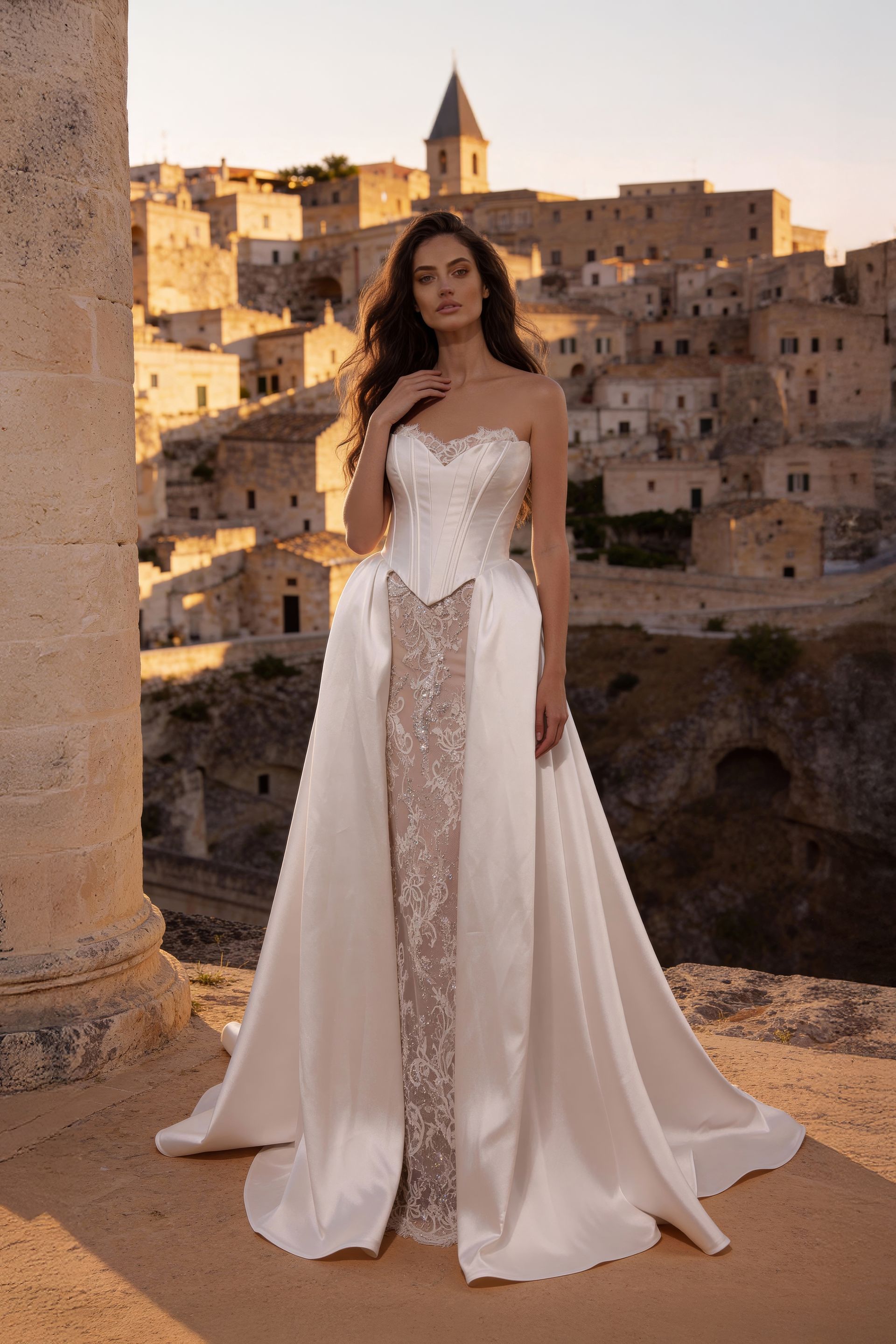 Woman in a white gown standing before a sunlit hillside village at sunset