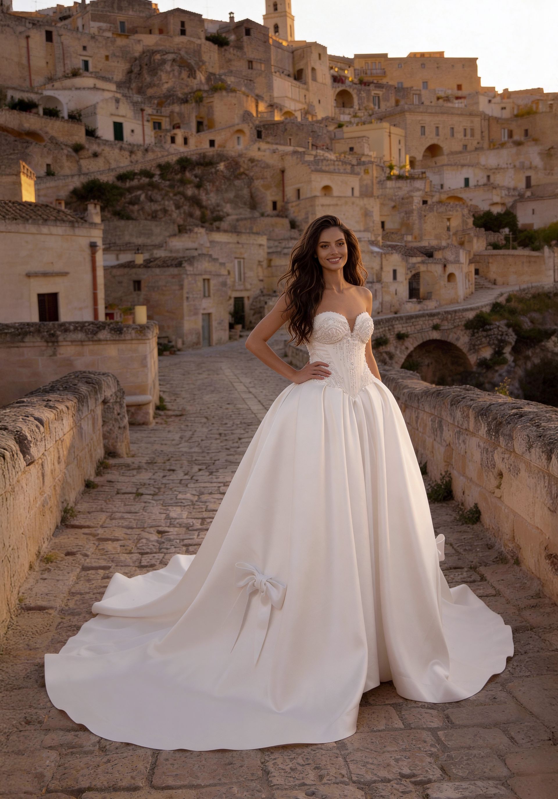 Bride in a white gown standing on a stone bridge in a historic hillside village at sunset