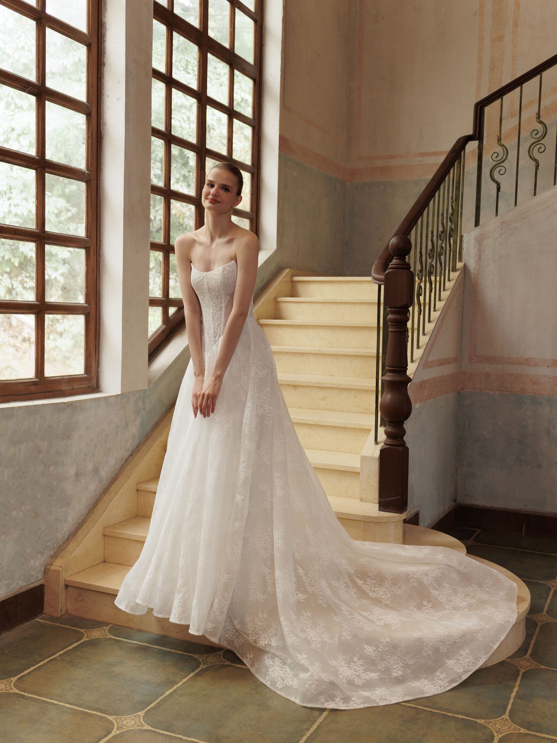 Woman in a white lace wedding gown standing by a staircase and tall window in a bright interior