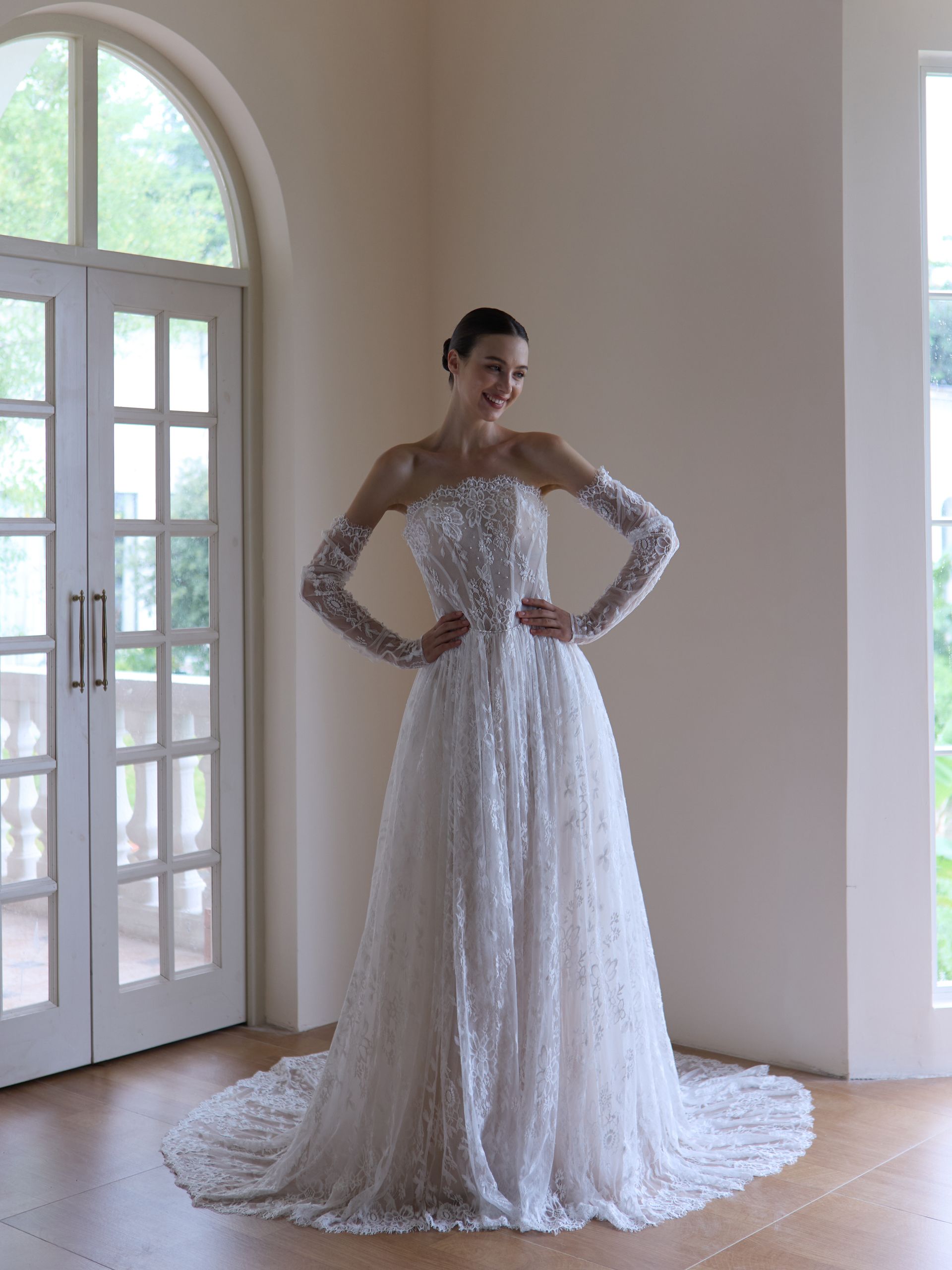 Person in a white lace gown standing in a bright room with hands on hips near tall windows