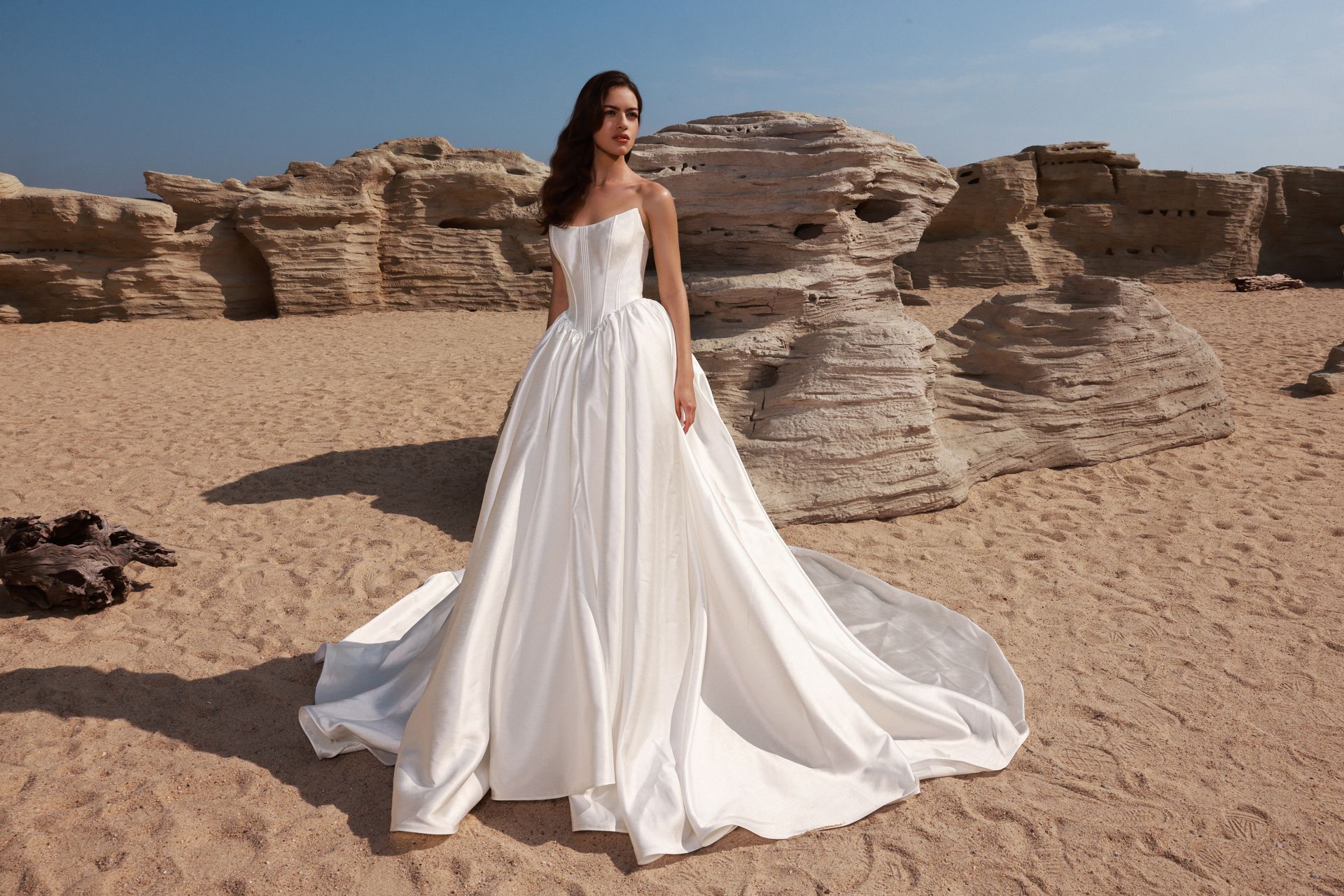 Woman in a white gown standing in a sandy desert with layered rock formations behind her.