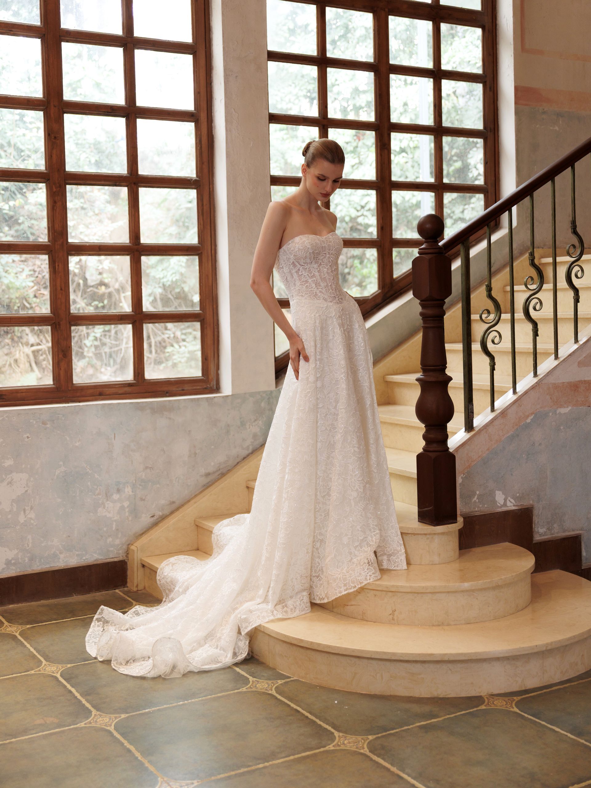 Bride in a strapless white lace gown standing on a staircase by large windows.