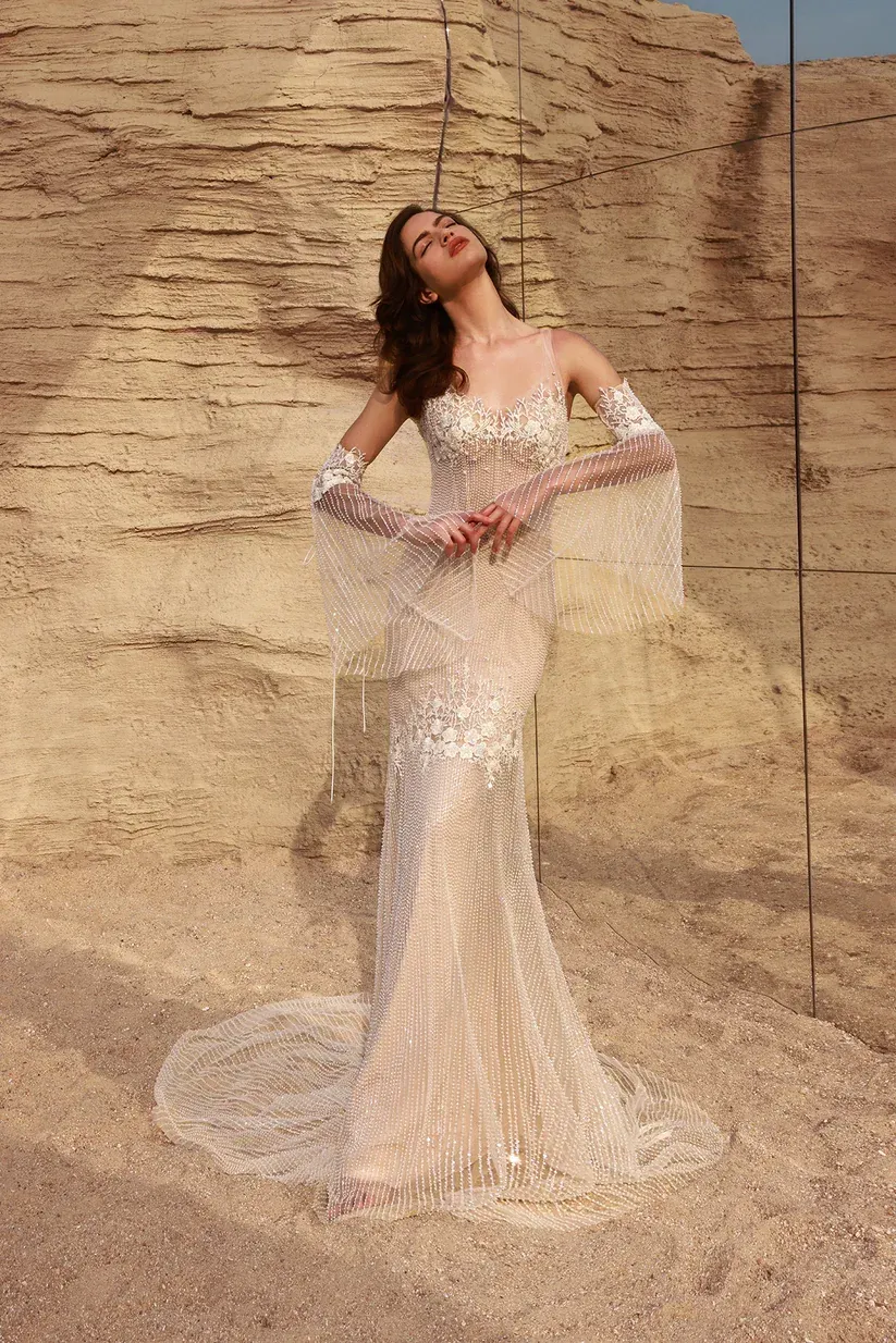 Woman in a white, embellished gown with long sleeves, posing outdoors against a rocky backdrop.