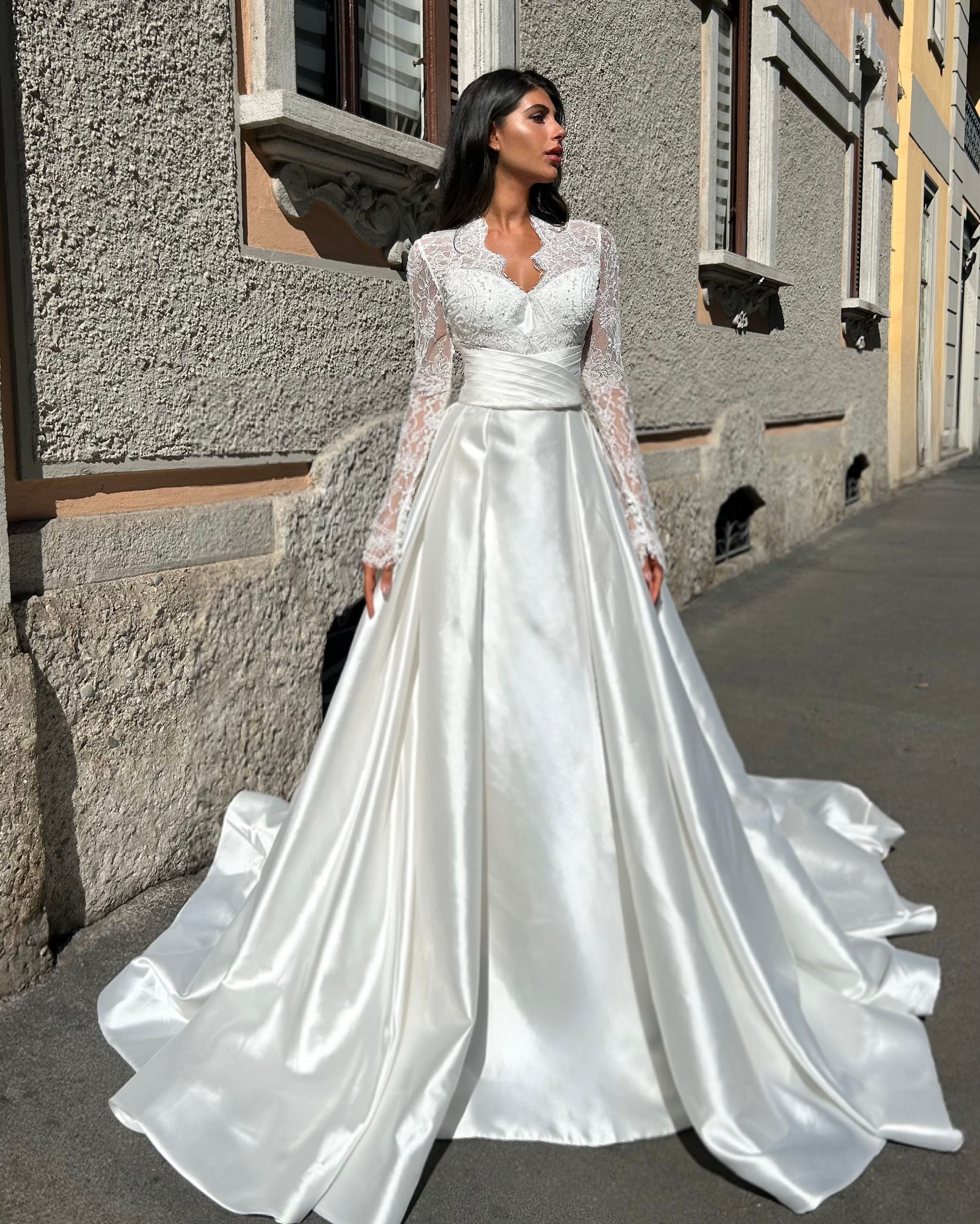 Woman in a white satin wedding gown posing outdoors beside a stone building