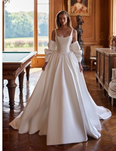 Woman in a white wedding gown standing in an elegant room with large windows and wooden furniture