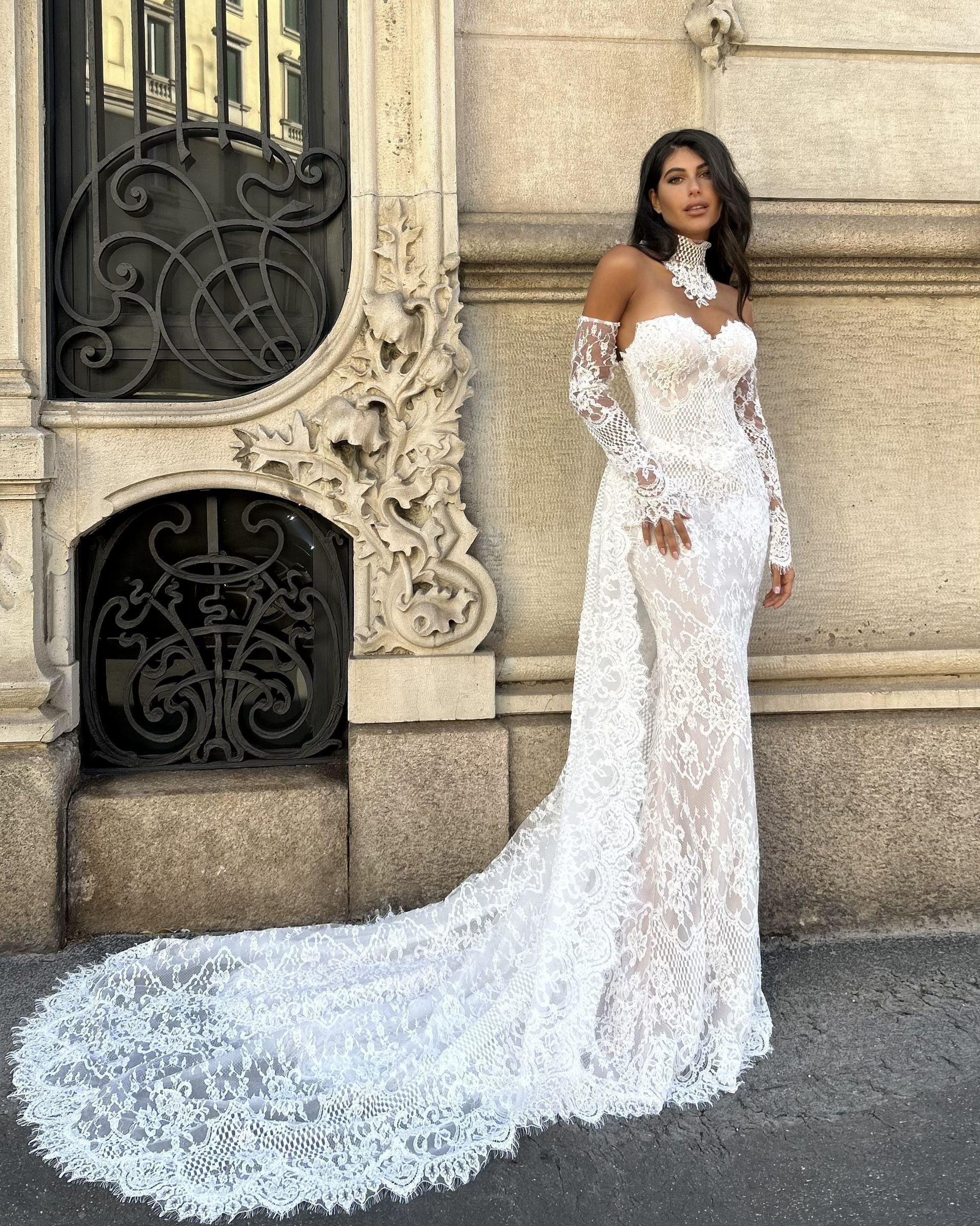 Woman in a white lace gown with a long train standing against an ornate stone wall.
