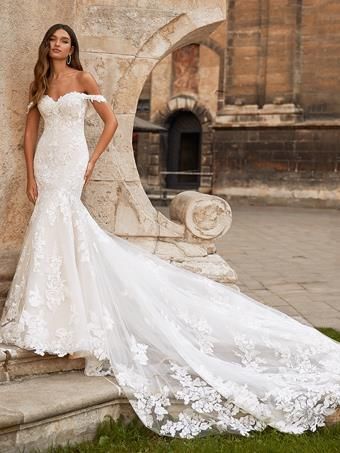 Woman in a white off-the-shoulder lace wedding dress, posed by stone archway, train trailing.