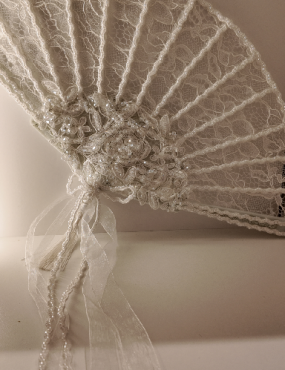 White lace decorative fan with sheer ribbon hanging against a beige wall
