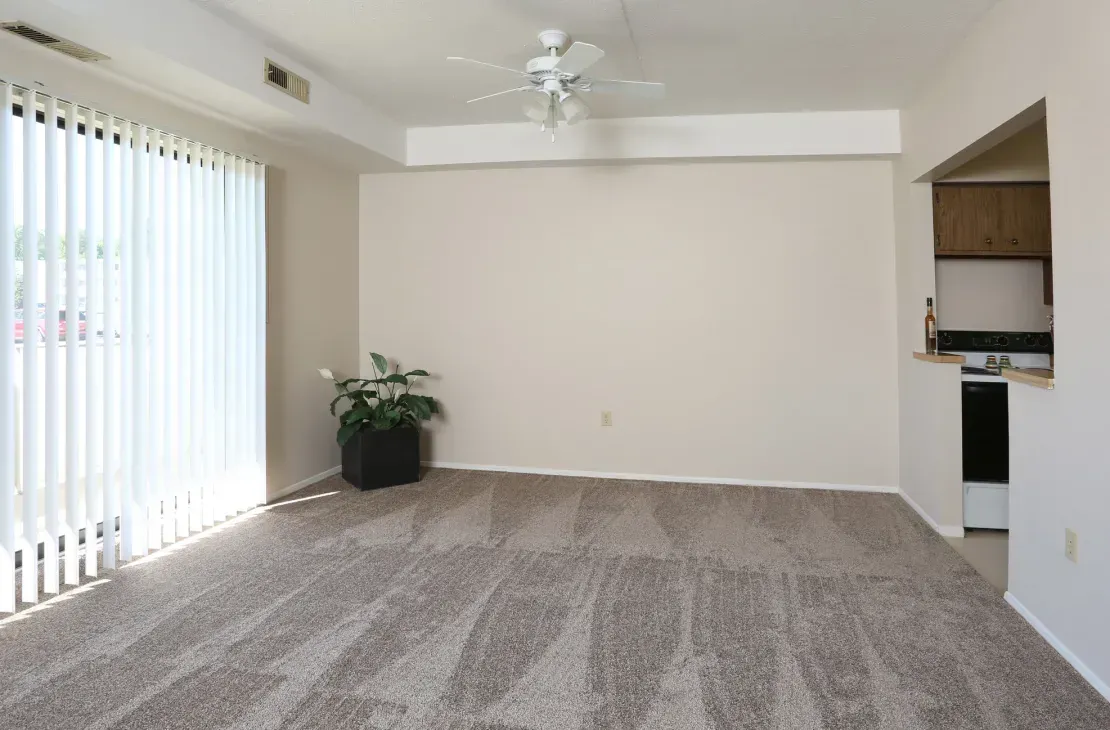 An empty living room with a ceiling fan and sliding glass doors leading to a kitchen.