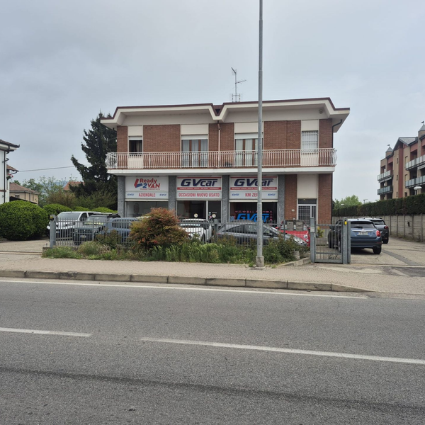 Edificio a due piani con vetrine, auto parcheggiate e una strada antistante sotto un cielo nuvoloso.