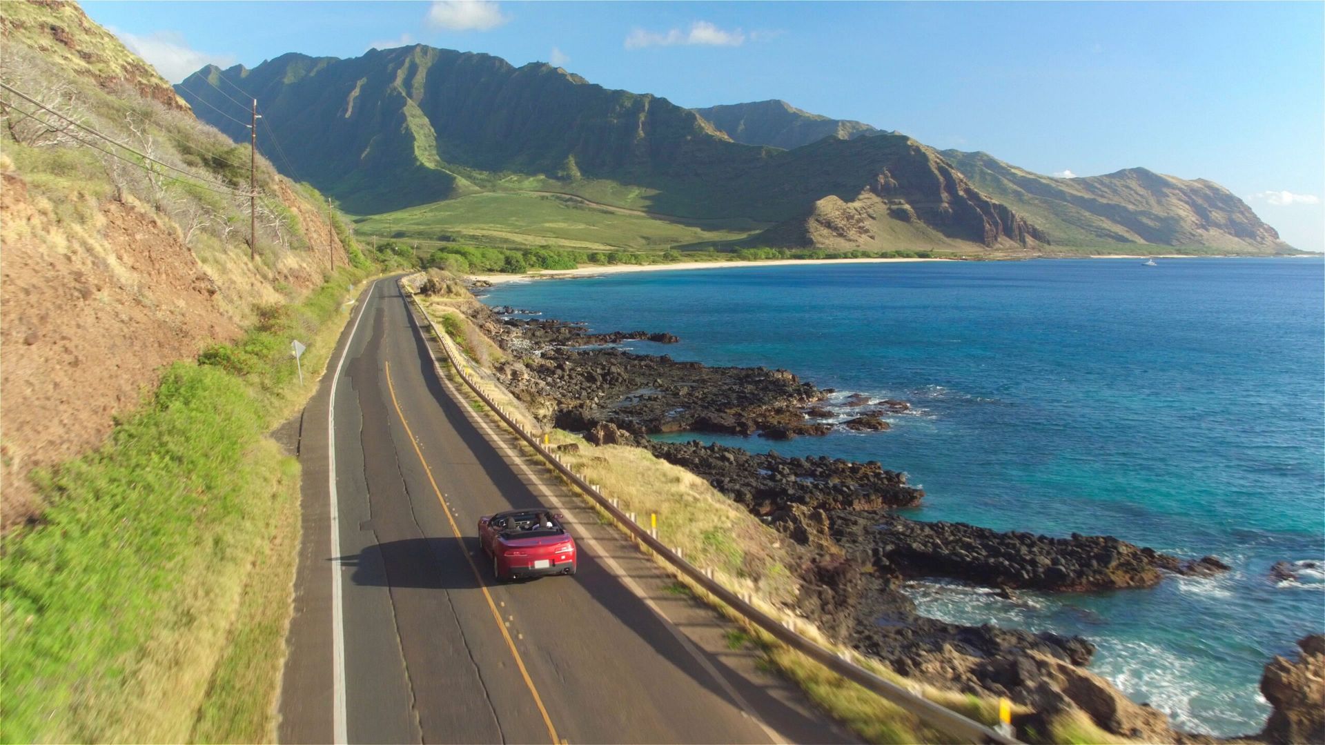 Red car driving along a coastal highway in front of mountains and turquoise ocean on a sunny day after being transported from Florida thanks to Budget Auto Transporter