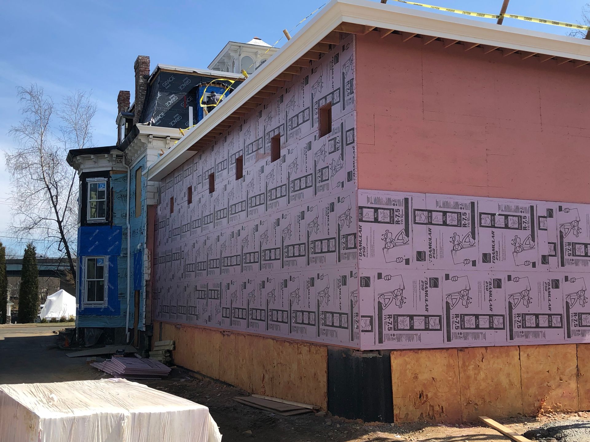 Construction site with pink insulation on a new building addition next to a blue-painted older building.