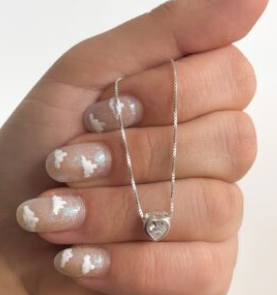 Mano sosteniendo un collar de corazón plateado sobre esmalte de uñas gris brillante con detalles florales blancos.
