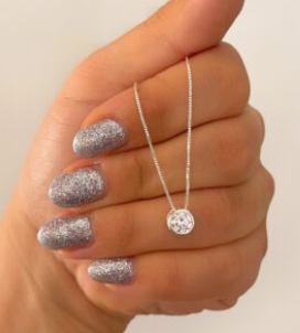 Mano con uñas plateadas brillantes sosteniendo un delicado collar de plata con un colgante redondo.