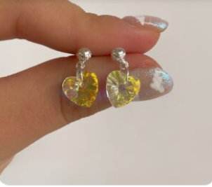 Una mano sostiene unos pendientes colgantes transparentes en forma de corazón con centros iridiscentes amarillos.