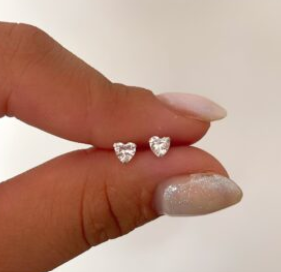Una mano sosteniendo dos pequeños pendientes de plata con forma de corazón sobre un fondo blanco.