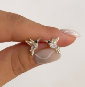 Pequeños pendientes de oro con forma de colibrí sostenidos entre los dedos sobre un fondo blanco.