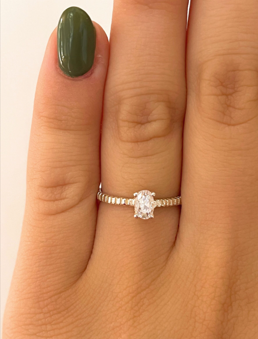 Mano con las uñas pintadas de verde oscuro, luciendo un anillo de plata con una piedra ovalada transparente.