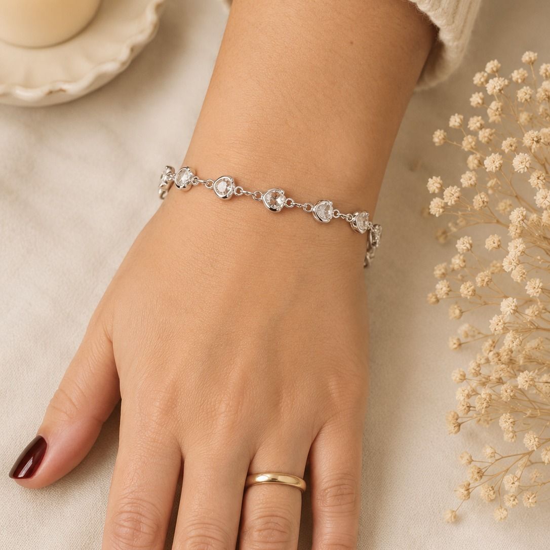 Mano con una pulsera floral plateada y un anillo dorado sobre un fondo beige.