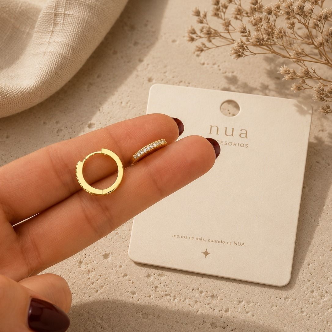 Una mano sostiene un pequeño pendiente de aro dorado junto a una tarjeta de exhibición de joyería beige.