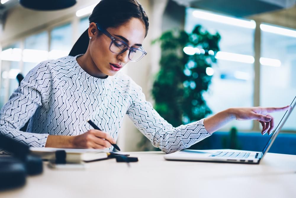 A Woman Is Sitting at A Desk Using a Laptop Computer and Writing in A Notebook — Sunshine Coast Financial Advisers in Maroochydore, QLD