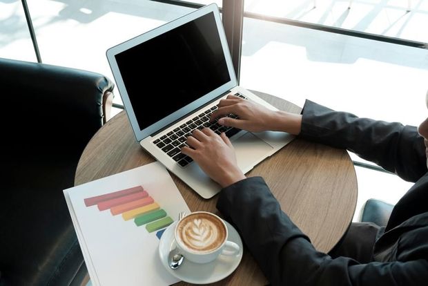 A Person Is Typing on A Laptop Computer While Sitting at A Table with A Cup of Coffee