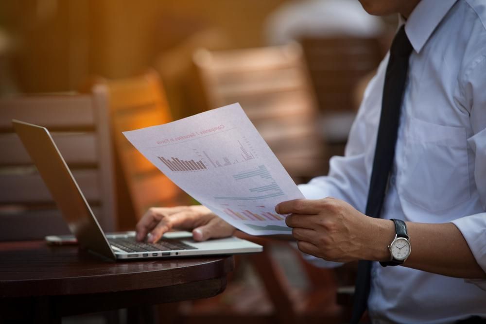 A Man Is Sitting at A Table Using a Laptop and Holding a Piece of Paper
