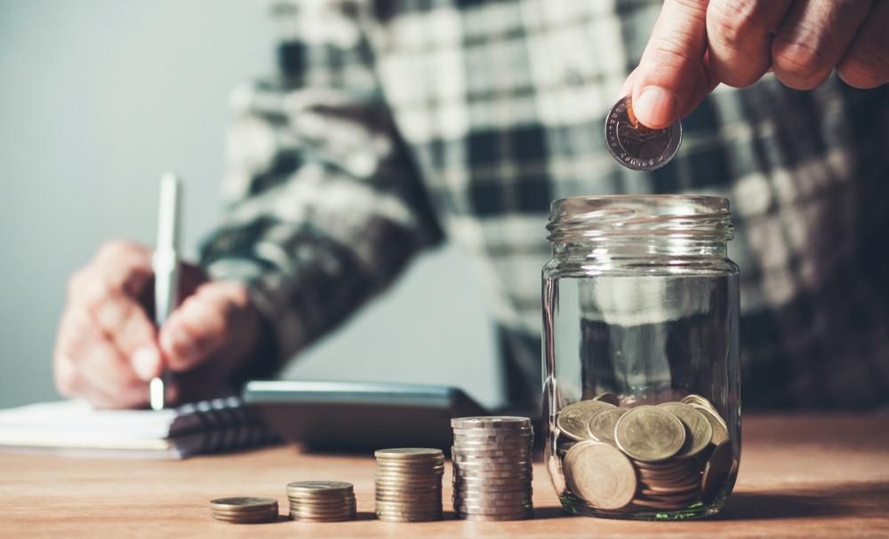 A Man Is Putting Coins Into a Jar While Writing in A Notebook — Sunshine Coast Financial Advisers in Maroochydore, QLD