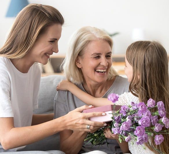 Two Women and A Little Girl Are Sitting on A Couch Holding Purple Flowers and A Gift Box