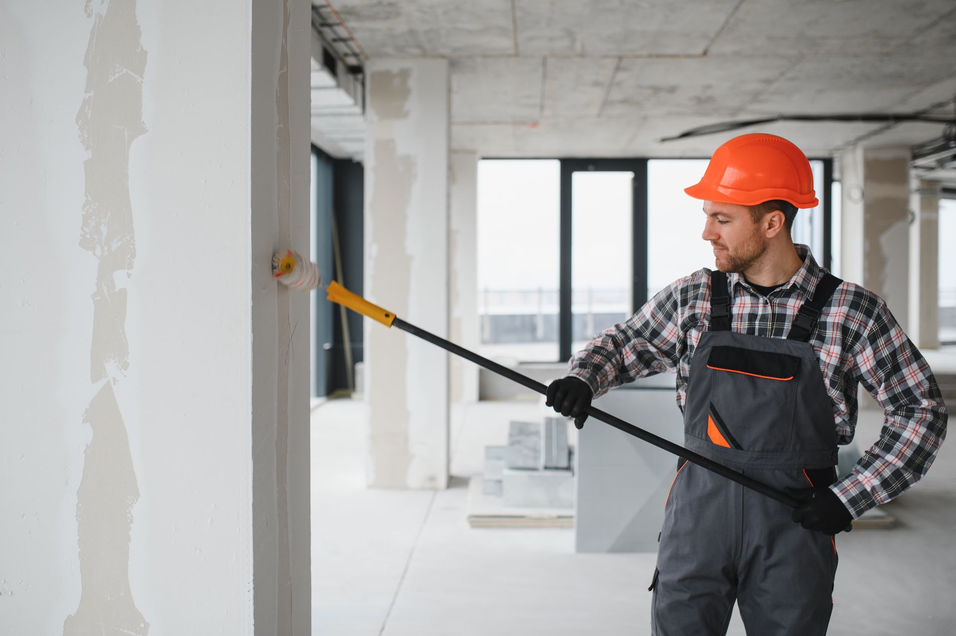 Worker in orange hard hat painting a wall in a bright, unfinished room