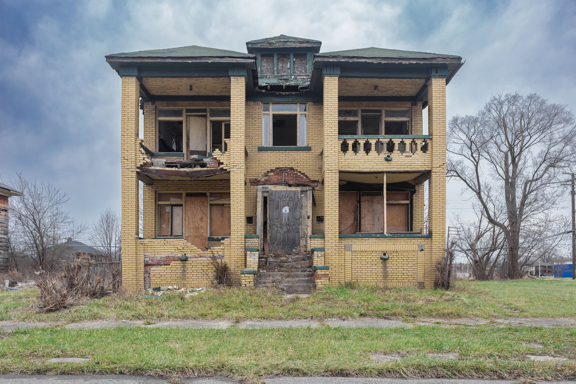 Abandoned two-story brick house with broken windows and overgrown grass under a cloudy sky