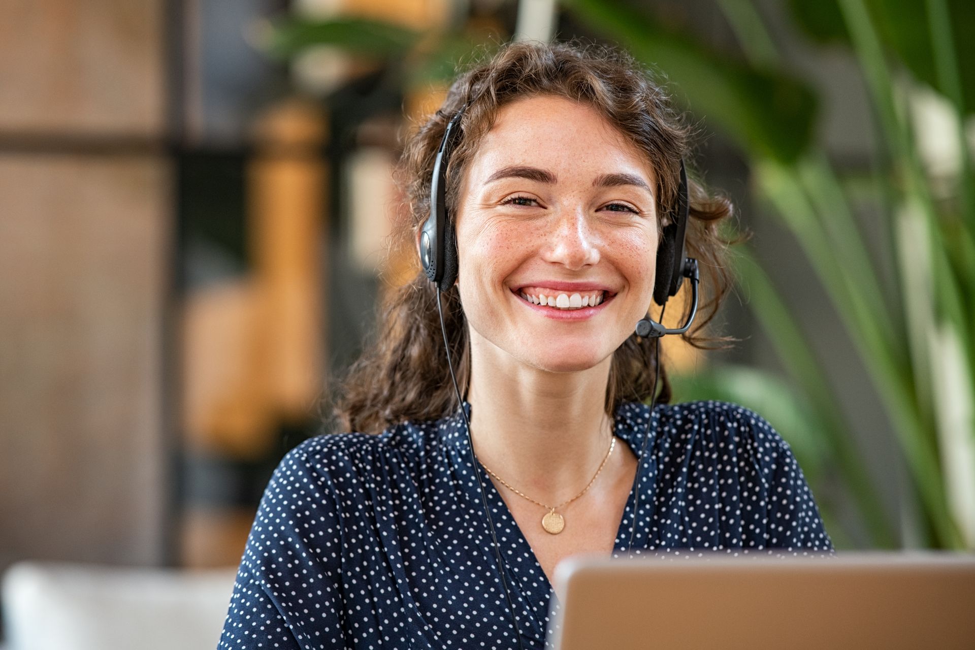 Customer support representative smiles while wearing a headset. She's in an office setting near a laptop and plants.