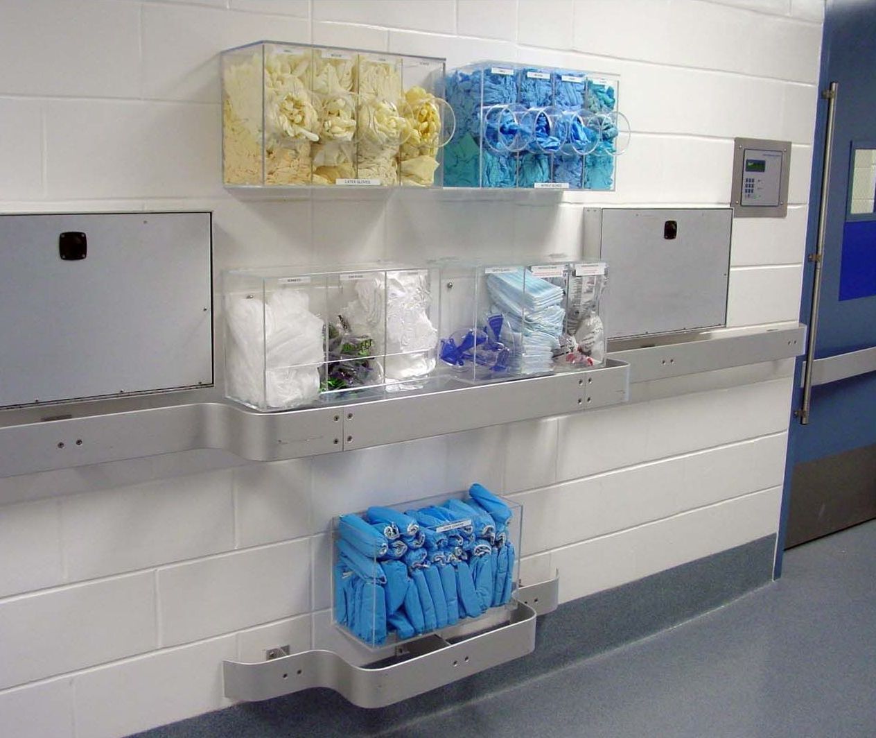 Clear PPE dispensers mounted to wall filled with various PPE masks, hairnets, and shoe covers.