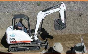 A man is driving a small excavator on a dirt road.