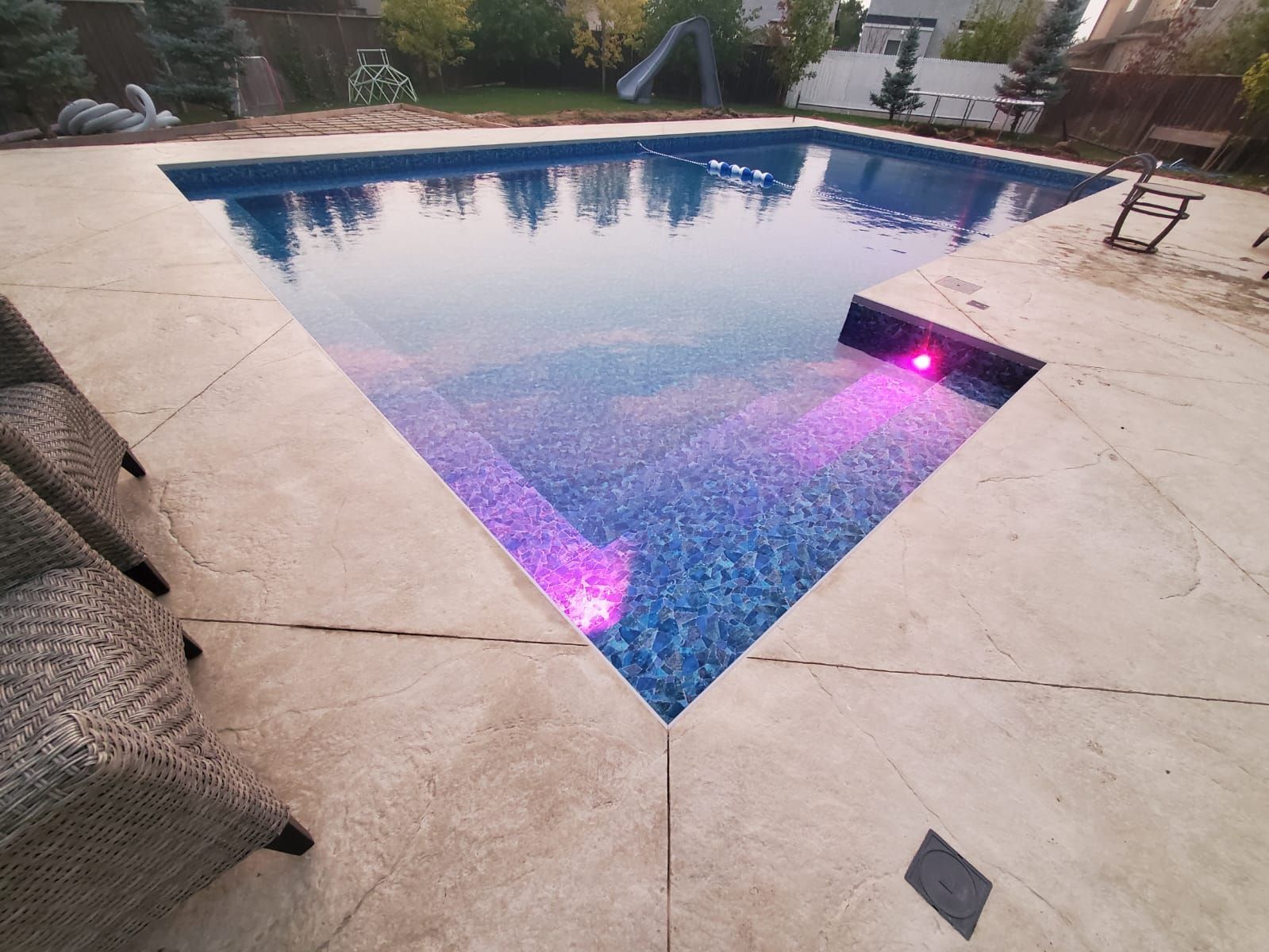 A rectangular backyard swimming pool with light-colored concrete decking, glowing purple underwater lights, and stairs.