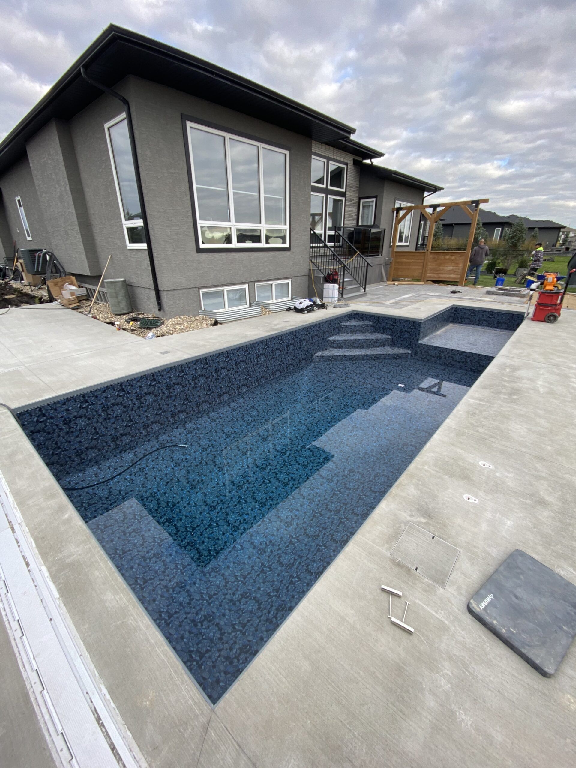 A modern house with gray stucco siding and a rectangular inground pool featuring dark blue patterned tiles and stairs.