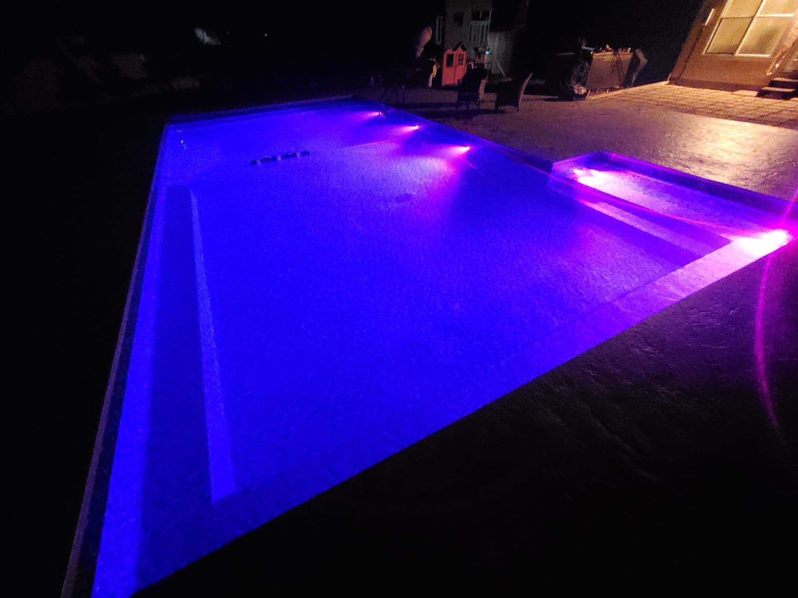 A backyard swimming pool at night, illuminated by vibrant blue and purple LED lights.