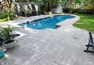 A backyard swimming pool with a gray stone patio, a small potted palm, and a wooden pergola in a lush green setting.