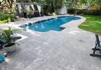 A backyard swimming pool with a gray stone patio, a small potted palm, and a wooden pergola in a lush green setting.