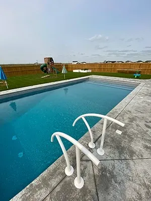 A rectangular backyard swimming pool with white handrails in the foreground, adjacent to a patio and grassy lawn.