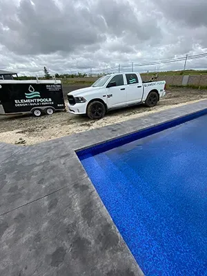 A white pickup truck and branded trailer parked next to a blue tiled swimming pool under a cloudy sky.